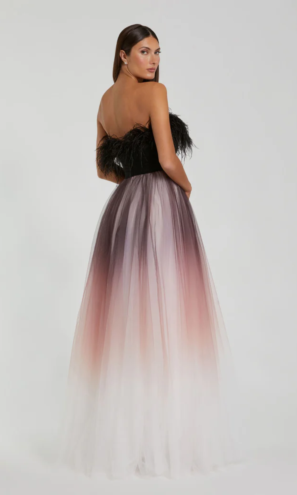 A woman stands with her back to the camera in the Long Formal Dress 20557 by Mac Duggal, a strapless ball gown with a black feathered bodice and ombre feather trim flowing into a long, A-line tulle skirt that fades from dark to light.